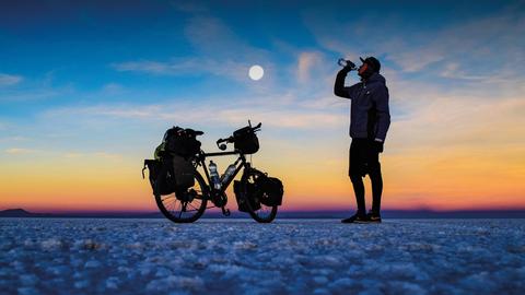 Szene aus der Doku "Besser Welt als nie": Dennis Kailing steht mit seinem Fahrrad in Boliviens Salar de Uyunineu und trinkt aus einer Wasserflasche.