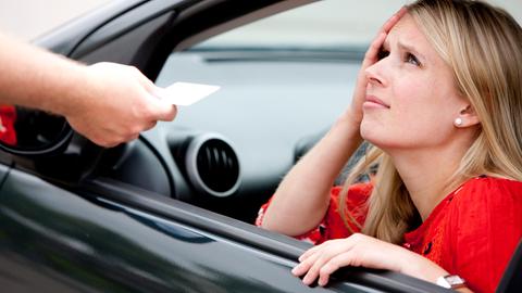 Eine Frau schaut verzweifelt aus ihrem Auto raus, weil ihr ein Bußgeldbescheid durchs Fenster gereicht wird.